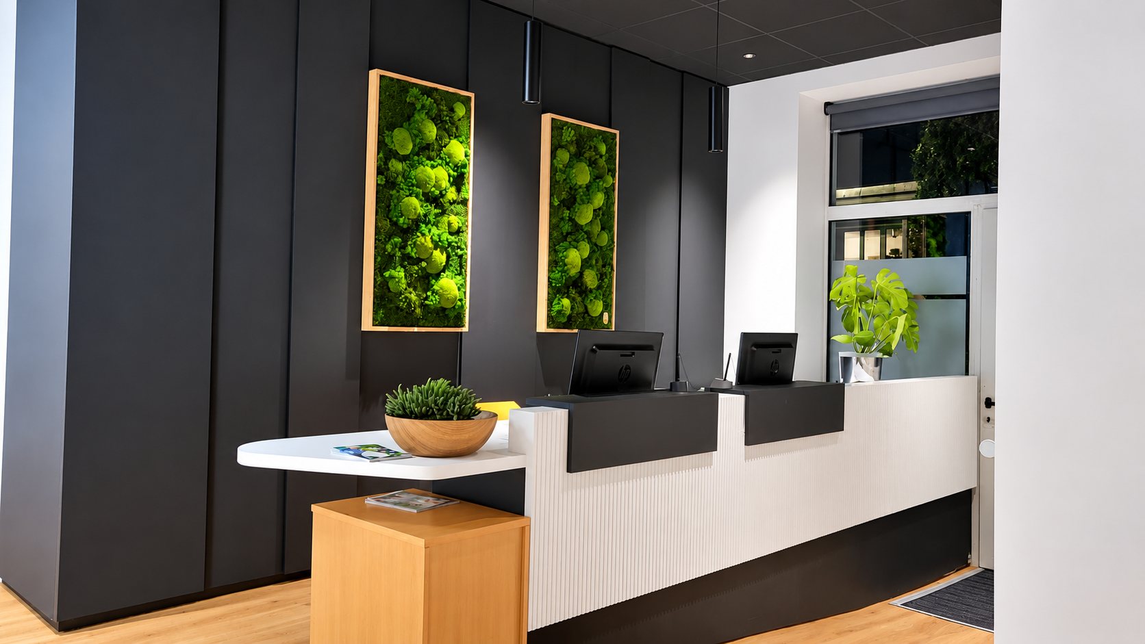 Un comptoir d'accueil blanc strié avec deux écrans d'ordinateur, devant un mur sombre orné de deux tableaux de mousse stabilisée encadrés de bois clair, avec des plantes vertes décoratives.