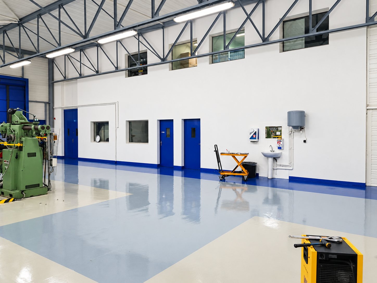 Intérieur d'un atelier industriel rénové avec un sol en résine brillante bleu et gris, des murs blancs, des portes bleues et une fraiseuse verte à gauche.