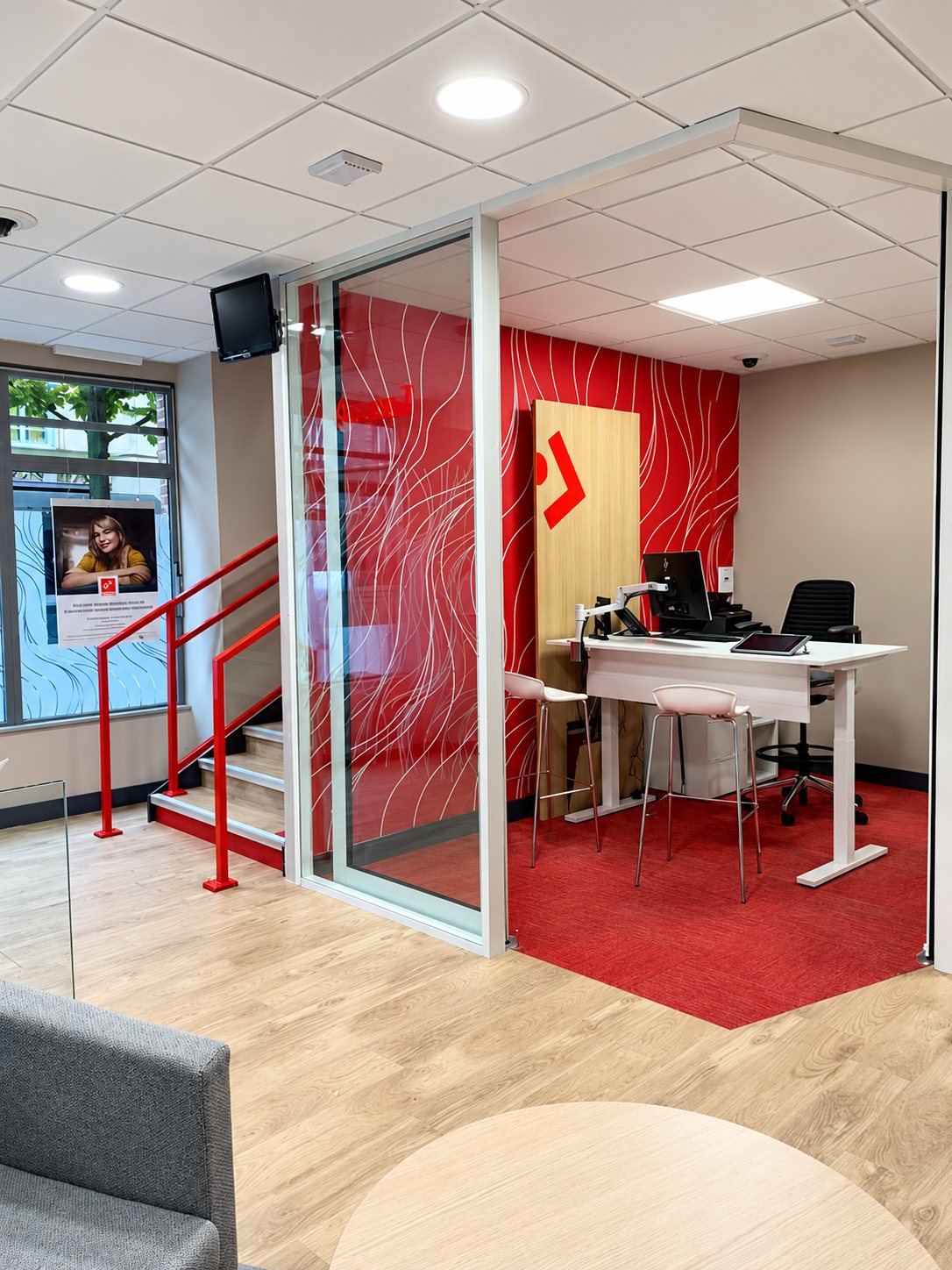Intérieur d'une agence aux tons rouges avec une cloison vitrée, un bureau réglable en hauteur, des tabourets hauts et un escalier à rambarde rouge.
