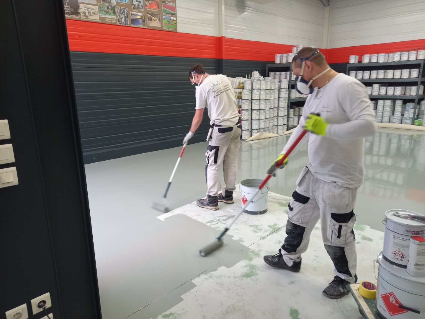 Deux ouvriers masqués appliquent au rouleau une résine grise sur le sol d'un entrepôt rempli de pots de peinture.