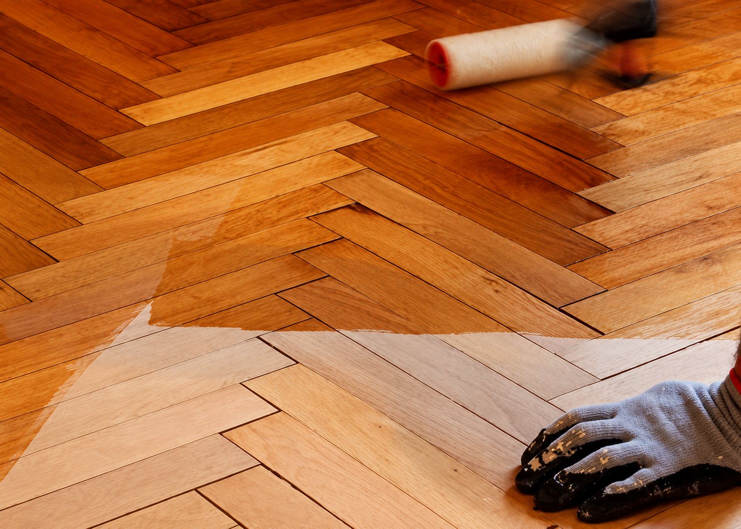 Un ouvrier ganté applique du vernis au rouleau sur un parquet en chevrons, la moitié du sol étant déjà traitée et plus foncée.
